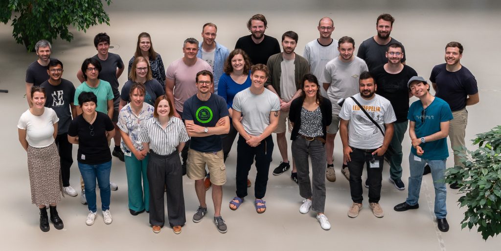 A group of people standing on a gray floor between two green plants, photographed from an angle slightly above.